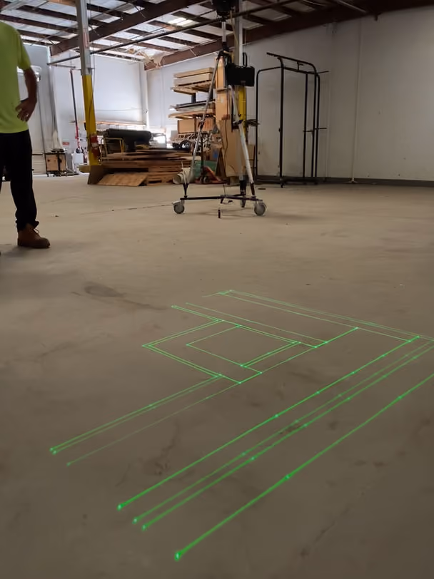 Green laser lines projecting drywall framing layout geometry onto a concrete floor from a digital layout tool, showing wall tracks and door opening positions at full scale