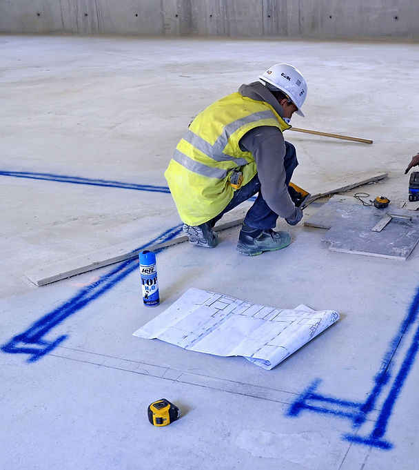 Construction worker manually marking drywall framing layout lines on a concrete floor using spray paint, with printed floor plans and tape measure beside him