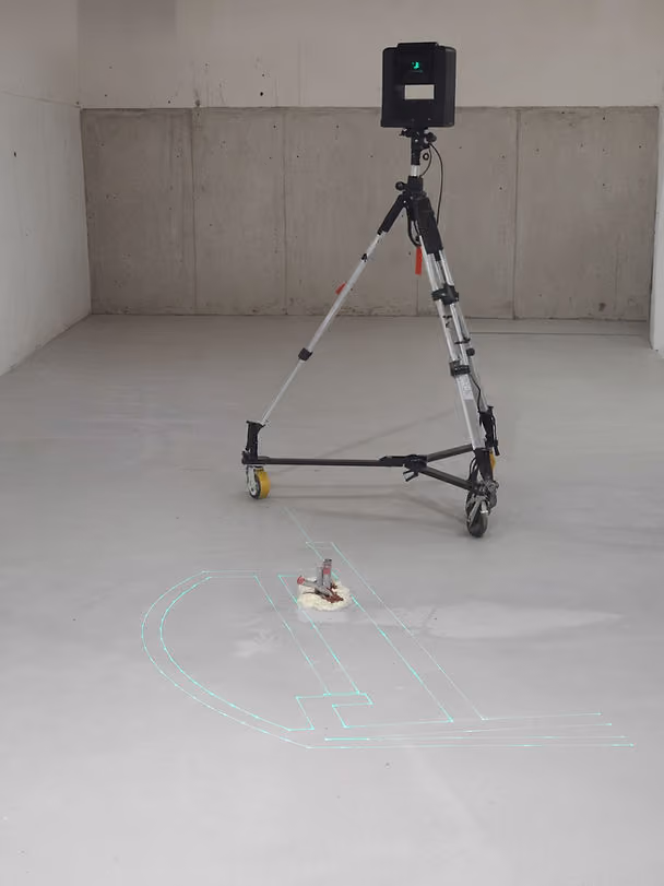A laser on a tripod projects green lines on a bare concrete floor in a minimalist room with white walls and a gray background.