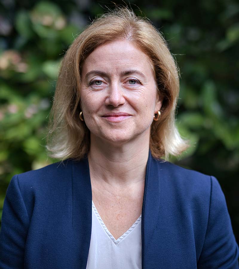 Smiling middle-aged woman with short blonde hair wearing a navy blazer and white blouse, standing outdoors with blurred greenery in the background.