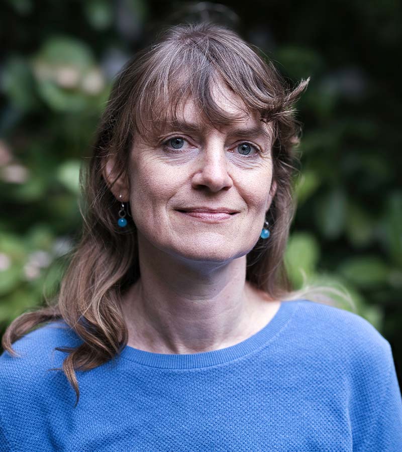Woman with light brown hair and blue eyes wearing a blue sweater and blue earrings in front of a blurred green foliage background.