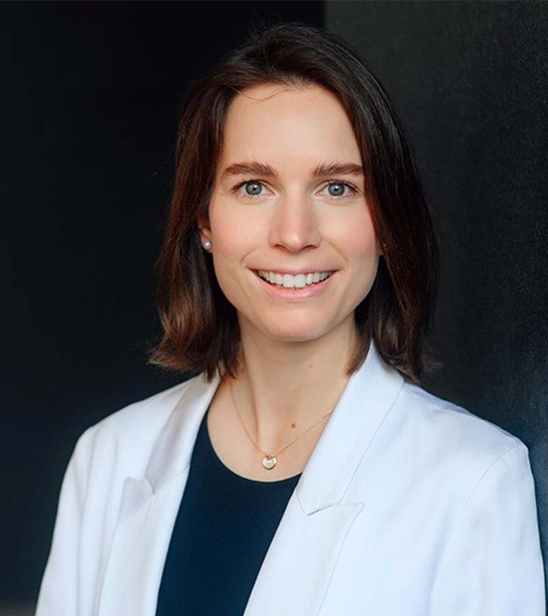 Smiling woman with shoulder-length brown hair wearing a white blazer, black top, pearl earrings, and a gold necklace against a dark background.