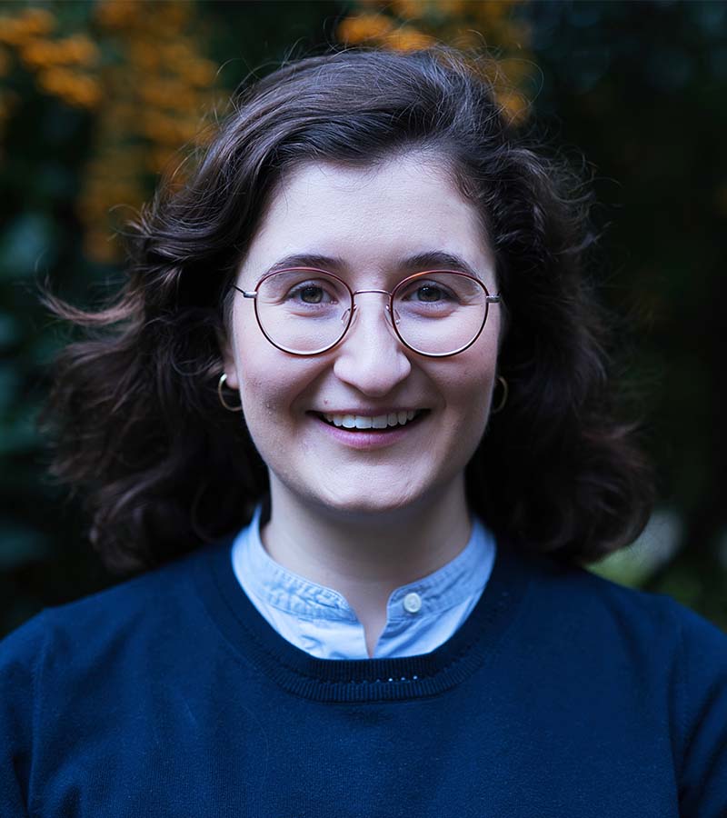 Smiling woman with wavy brown hair, round glasses, hoop earrings, wearing a blue shirt and dark sweater outdoors.