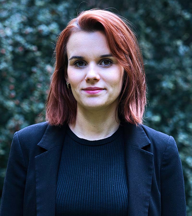 Portrait of a woman with shoulder-length red hair wearing a black blazer and top, standing outdoors with a blurred green foliage background.
