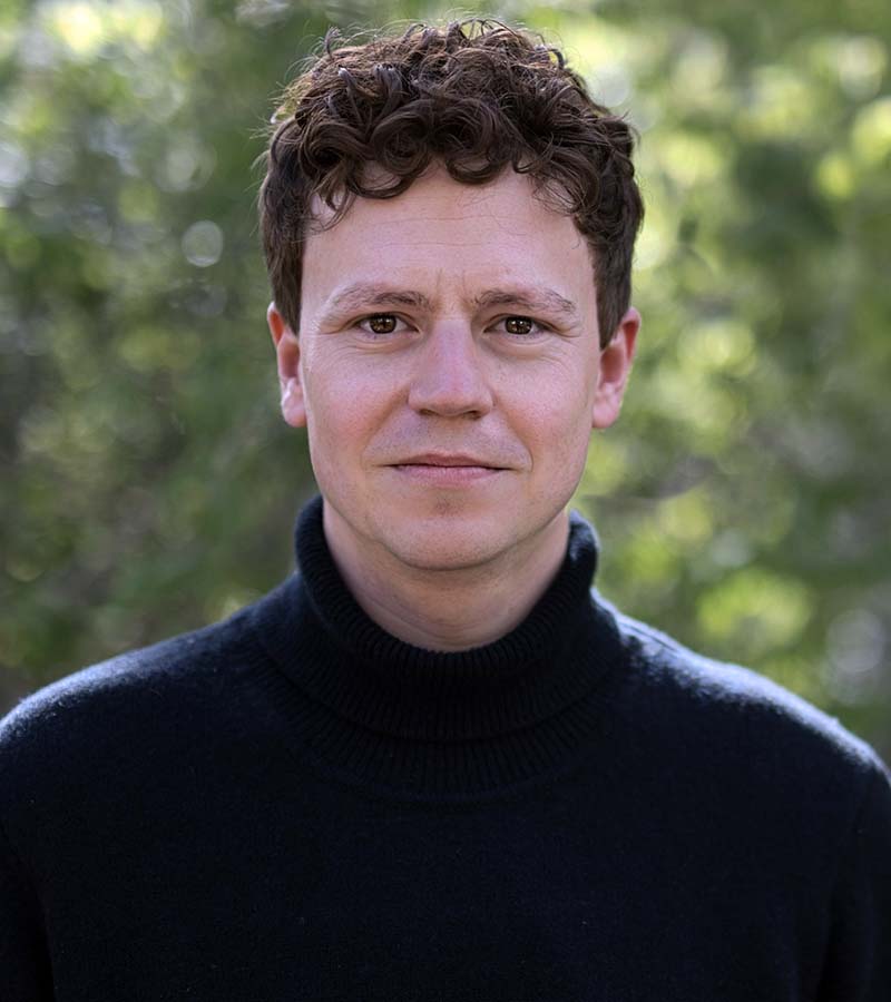 Man with curly brown hair wearing a black turtleneck sweater standing outdoors with blurred green foliage background.