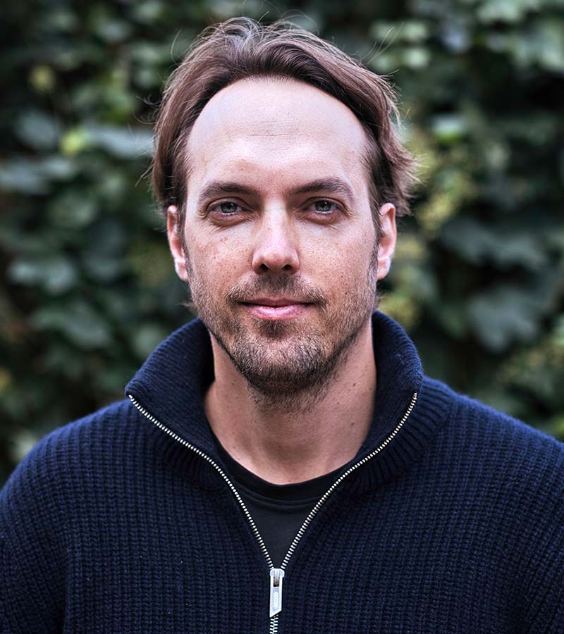 Portrait of a man with light brown hair and beard wearing a dark blue zip-up sweater, standing outdoors with blurred green foliage background.