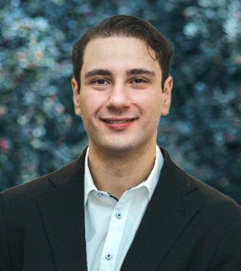Smiling young man with dark hair wearing a black suit jacket and white shirt against a blurred blue and green background.