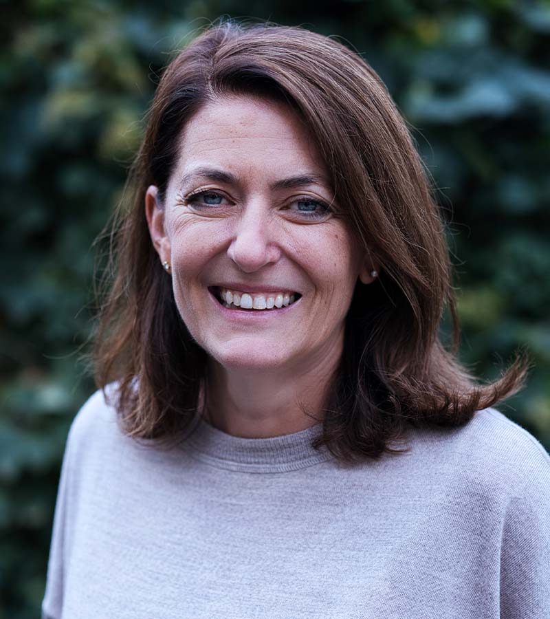 Smiling woman with shoulder-length brown hair wearing a light gray sweater, with a blurred background of green foliage.