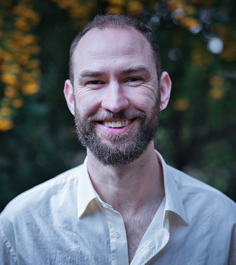 Smiling man with short hair and beard wearing a white shirt outdoors with blurred green and yellow foliage background.
