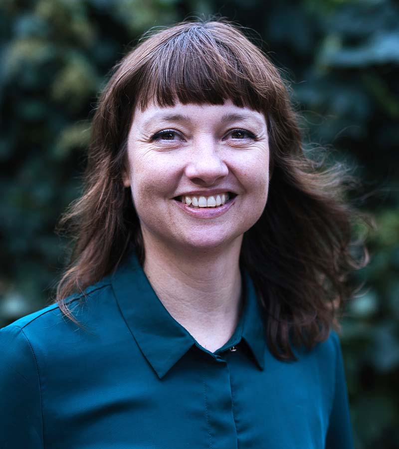 Smiling woman with medium-length brown hair and bangs wearing a teal collared shirt, with a blurred green foliage background.