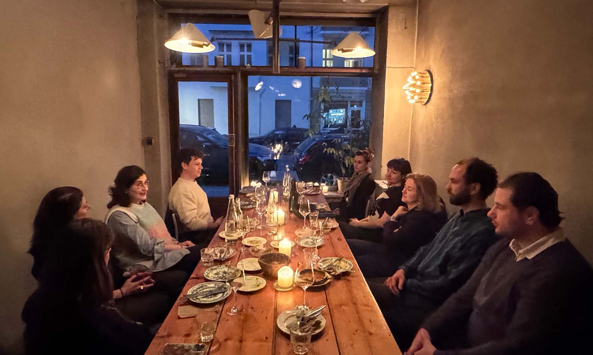 Group of eight people sitting around a long wooden table with plates, glasses, and candles in a warmly lit room.