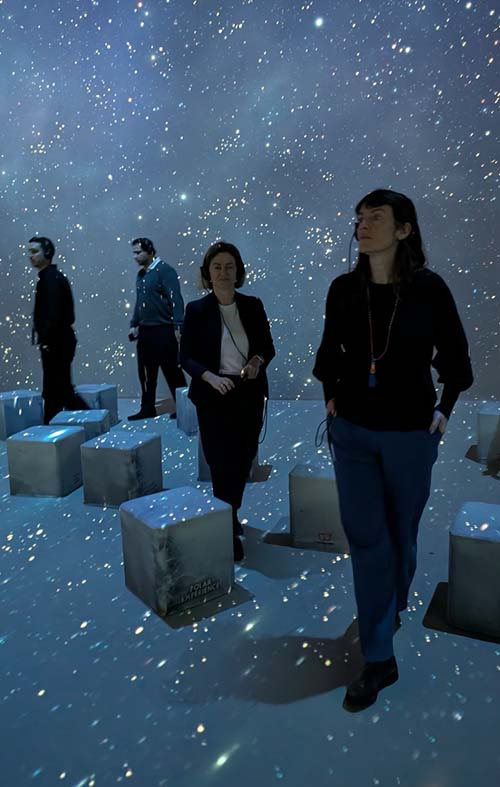 Four people standing and sitting among illuminated cube seats in a dark space filled with star-like light projections.
