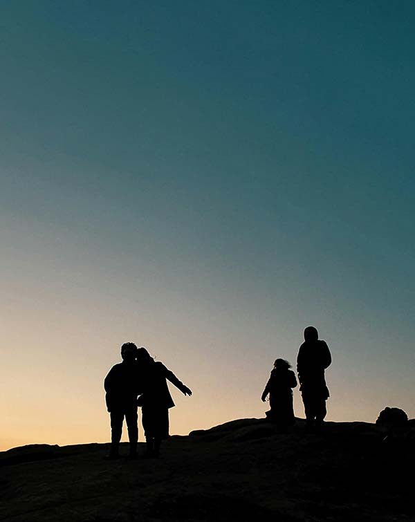 Silhouettes of four people on a hill during sunset with a gradient sky from pale yellow to deep blue.