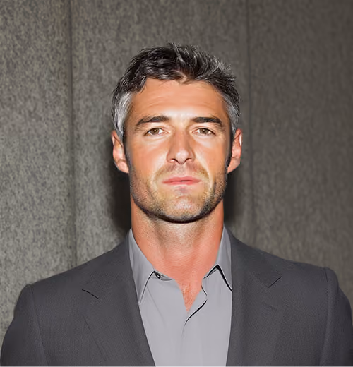 Portrait of a man with short salt-and-pepper hair wearing a gray shirt and dark blazer, standing in front of a stone wall.