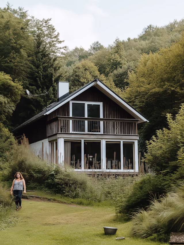 Two-story wooden house with large glass windows surrounded by dense green trees and shrubs, and a woman walking on a grass path nearby.