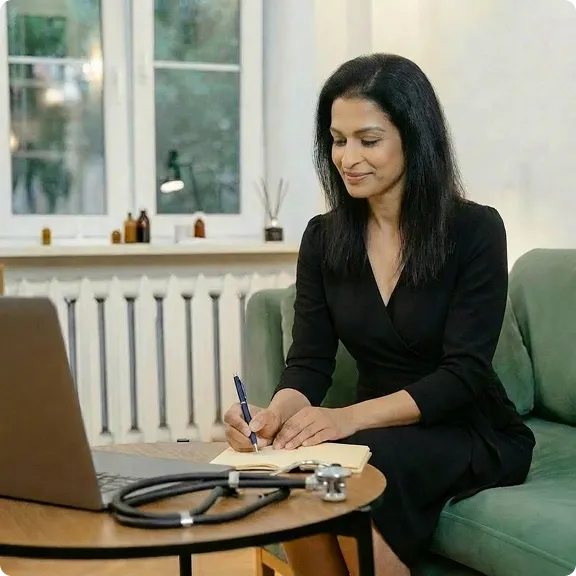 Woman in black dress sitting on green sofa, writing in a notebook with a laptop and stethoscope on the table in front of her.