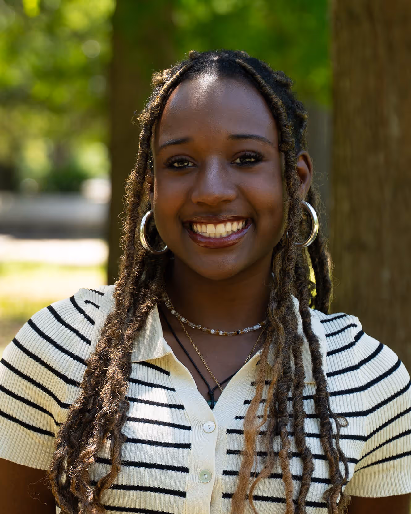 Smiling young woman with long braided hair wearing a striped shirt outdoors in sunlight.