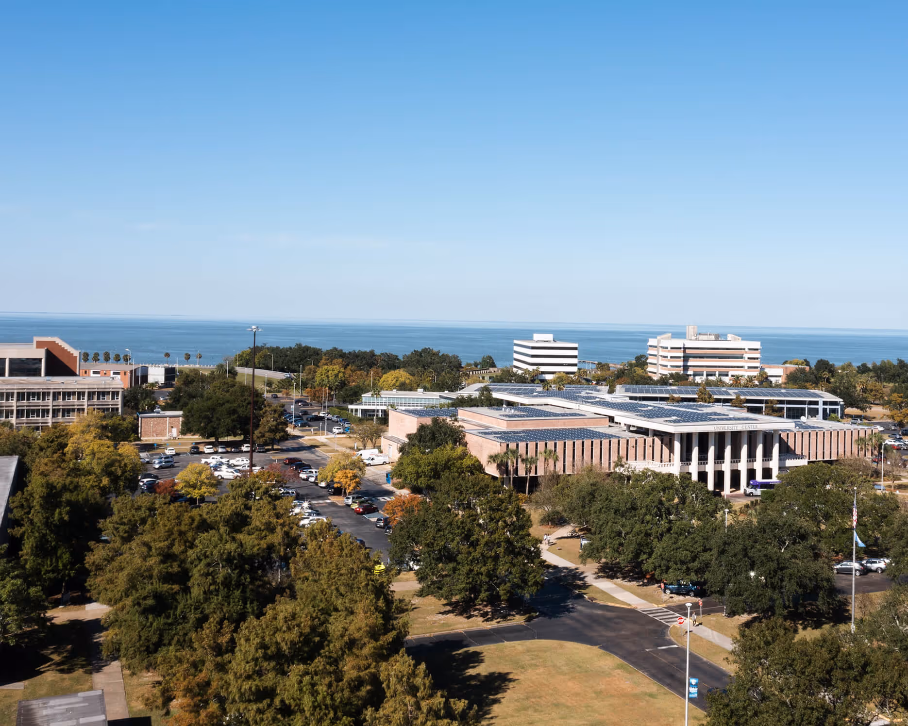 Wide aerial view of LSU New Orleans campus with buildings, parking lots, trees, and a large body of water in the background.