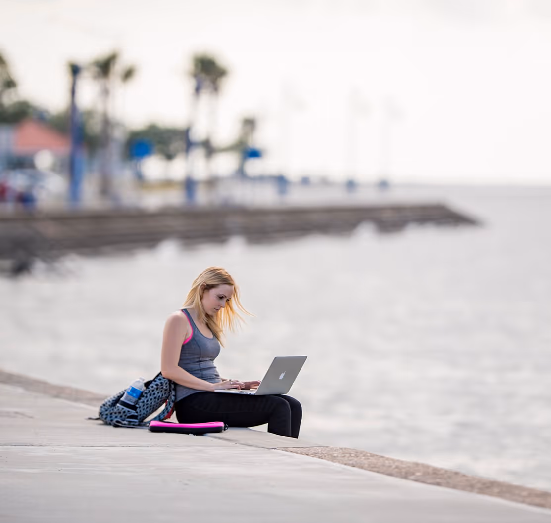 Young woman using a laptop sitting on a concrete ledge by the water with a backpack next to her.
