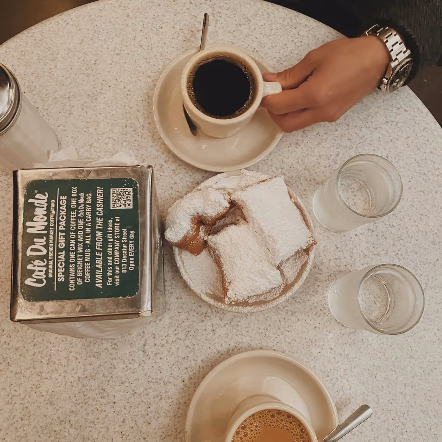 Table with two coffees, powdered sugar beignets on a plate, two glasses of water, and a hand holding a coffee cup.