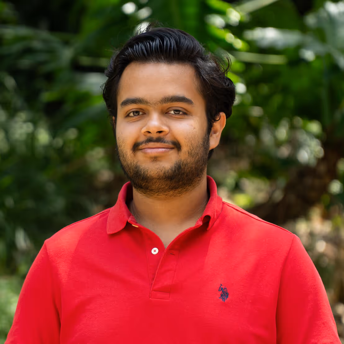 Smiling young man with dark hair and beard wearing a red polo shirt outdoors with green foliage background.