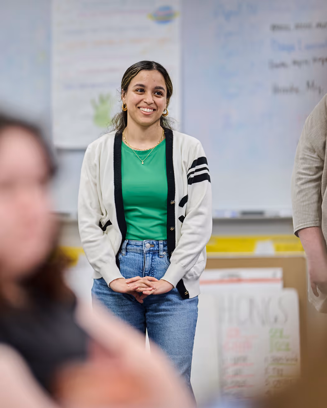 Smiling young woman in green shirt, white cardigan, and jeans standing in a classroom.