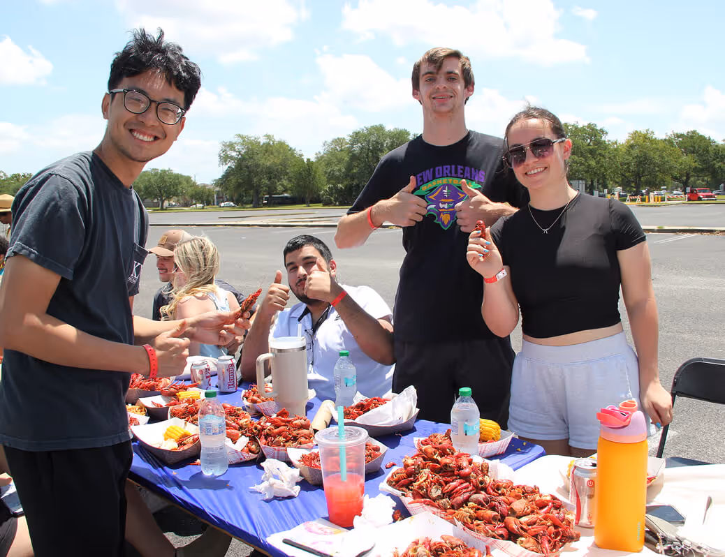 Four smiling young adults giving thumbs up around a table full of crawfish and drinks outdoors.