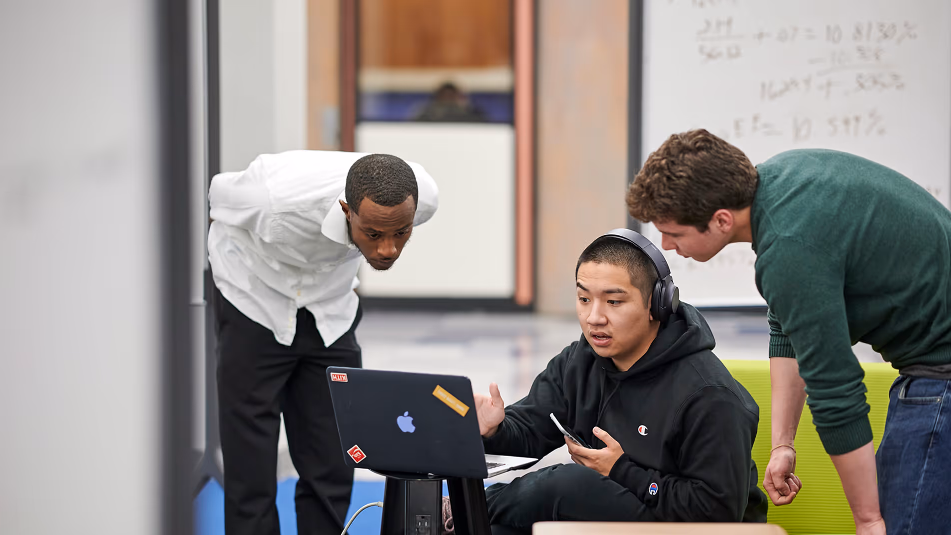 Three men focused on a laptop, one sitting wearing headphones, others standing and leaning in.
