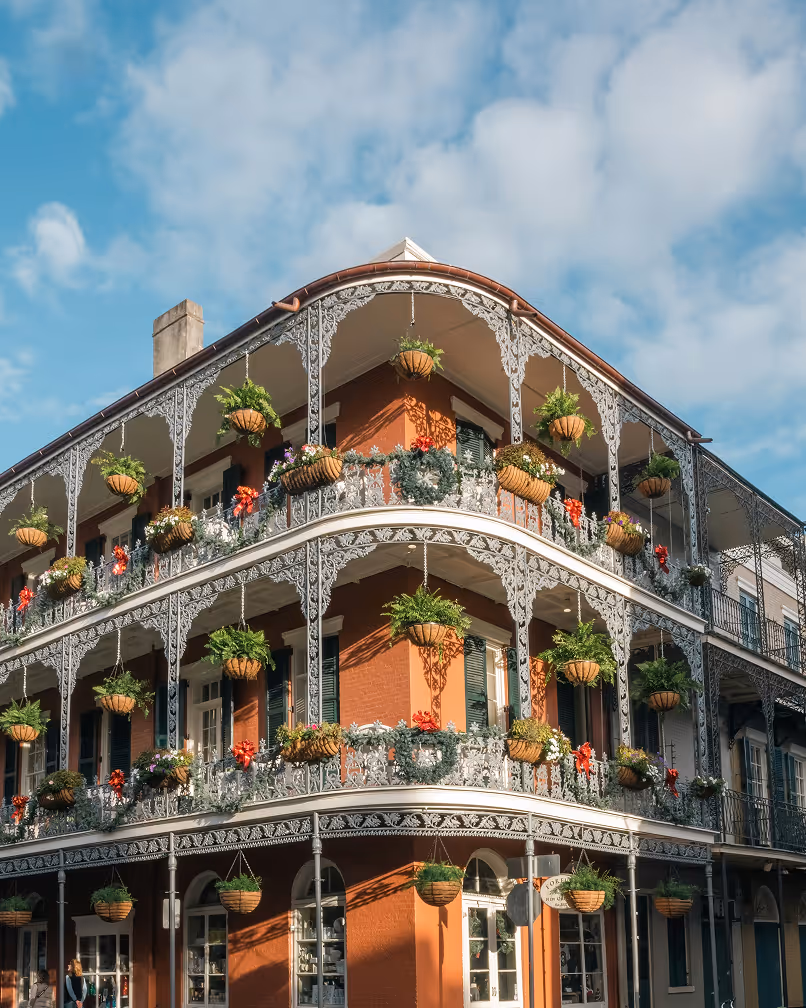 Ornate iron balconies with hanging plants and flowers on a corner building under blue sky.