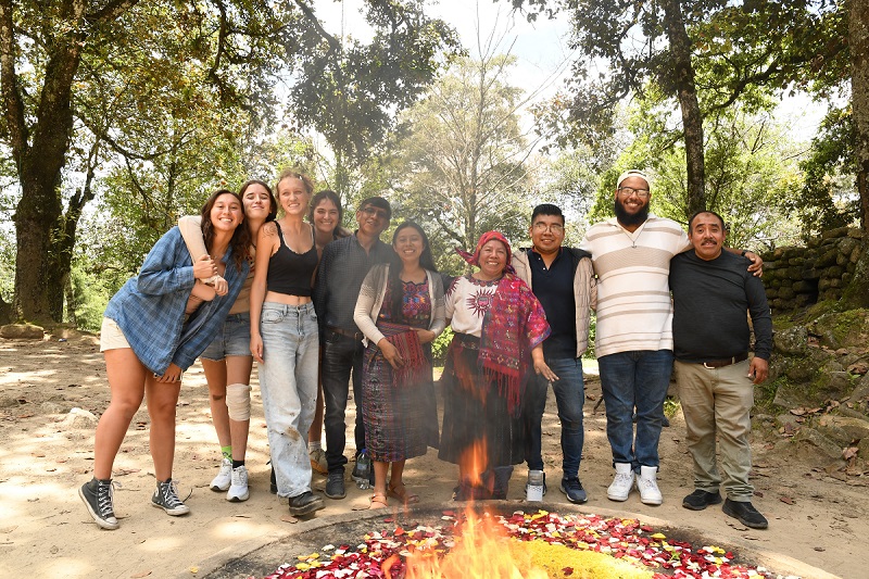 Group picture of students with locals in Guatemala surrounded by trees with a campfire in front