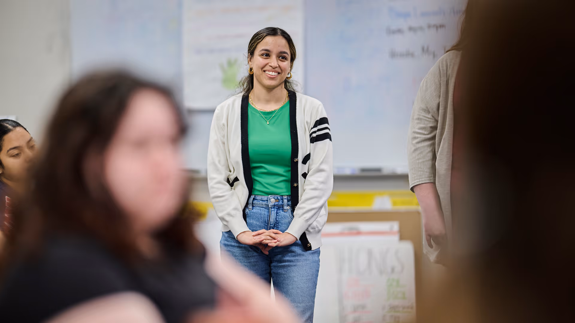 Young woman in green shirt and cardigan smiling and standing in a classroom with blurred people in foreground.