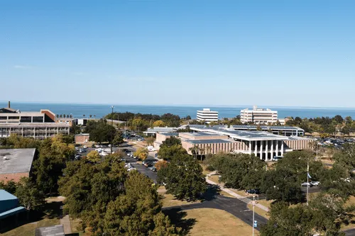 Wide aerial view of LSU New Orleans campus with buildings, parking lots, trees, and a large body of water in the background.