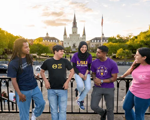 Five diverse students smiling and talking by railing with St. Louis Cathedral in the background at sunset.