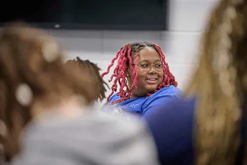 Person with pink dreadlocks and glasses smiling among a group in a casual indoor setting.
