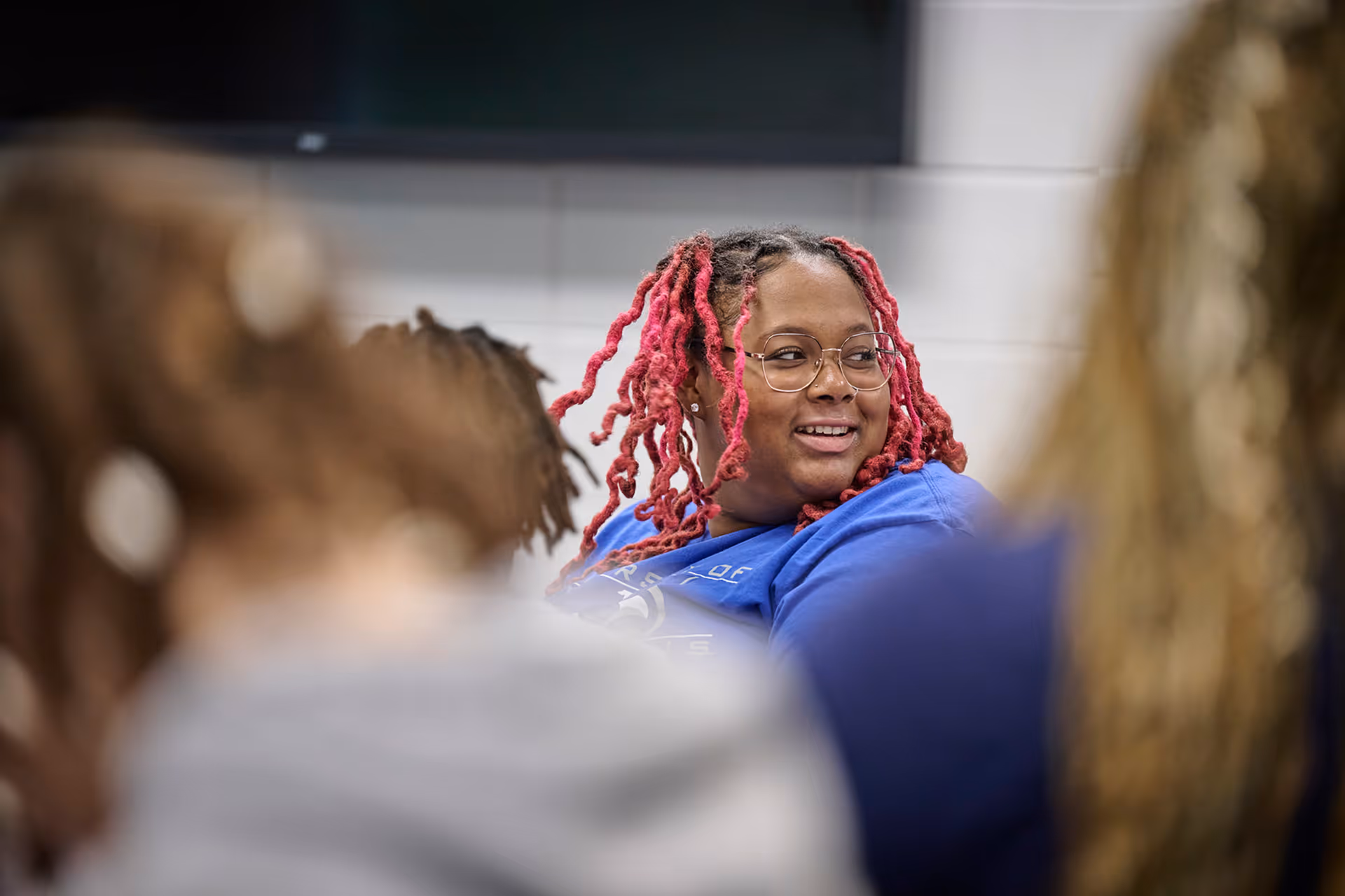 Person with pink dreadlocks and glasses smiling among a group in a casual indoor setting.
