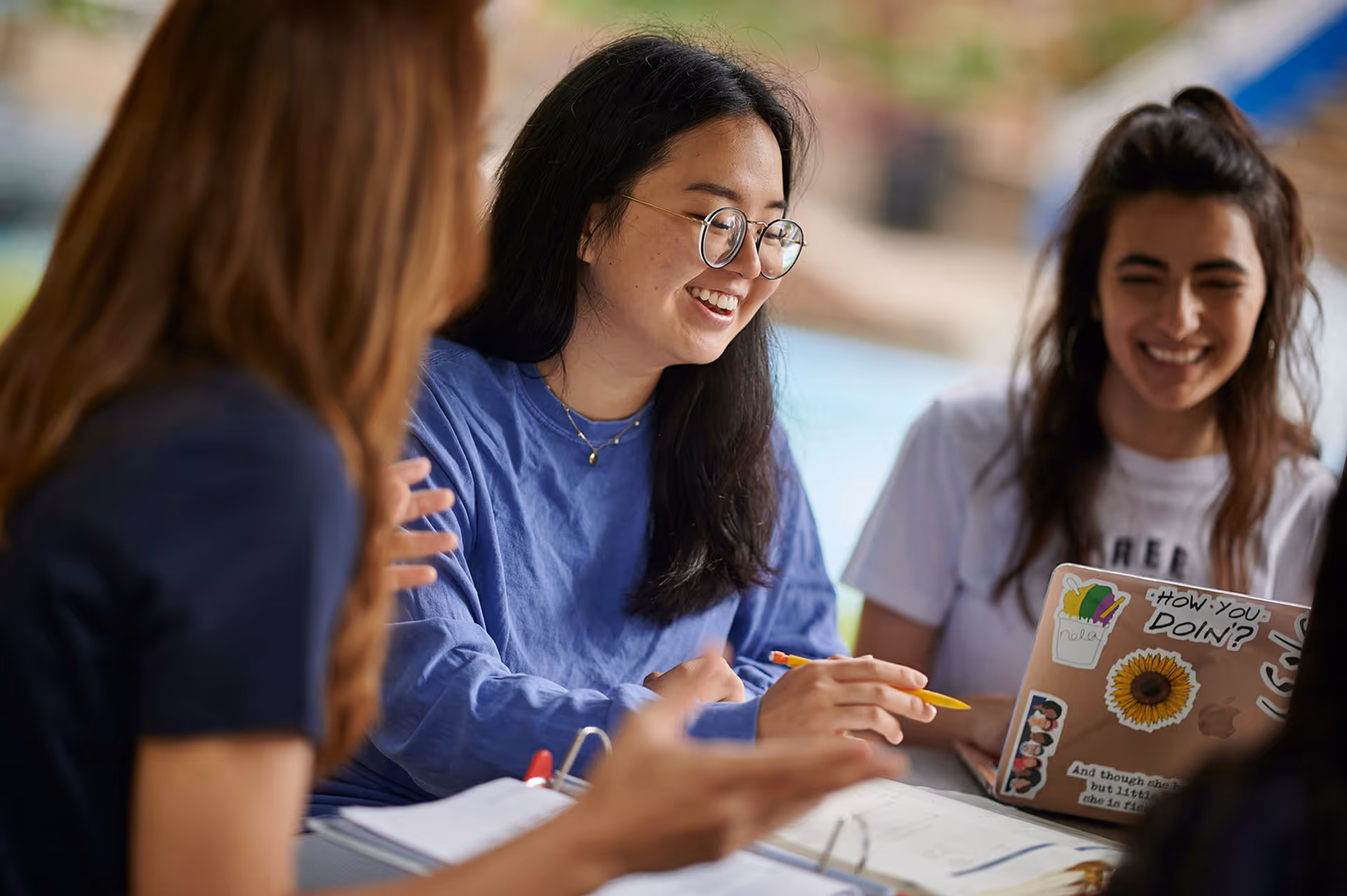 Three young women studying together, one pointing with a yellow pencil, smiling with a laptop open.