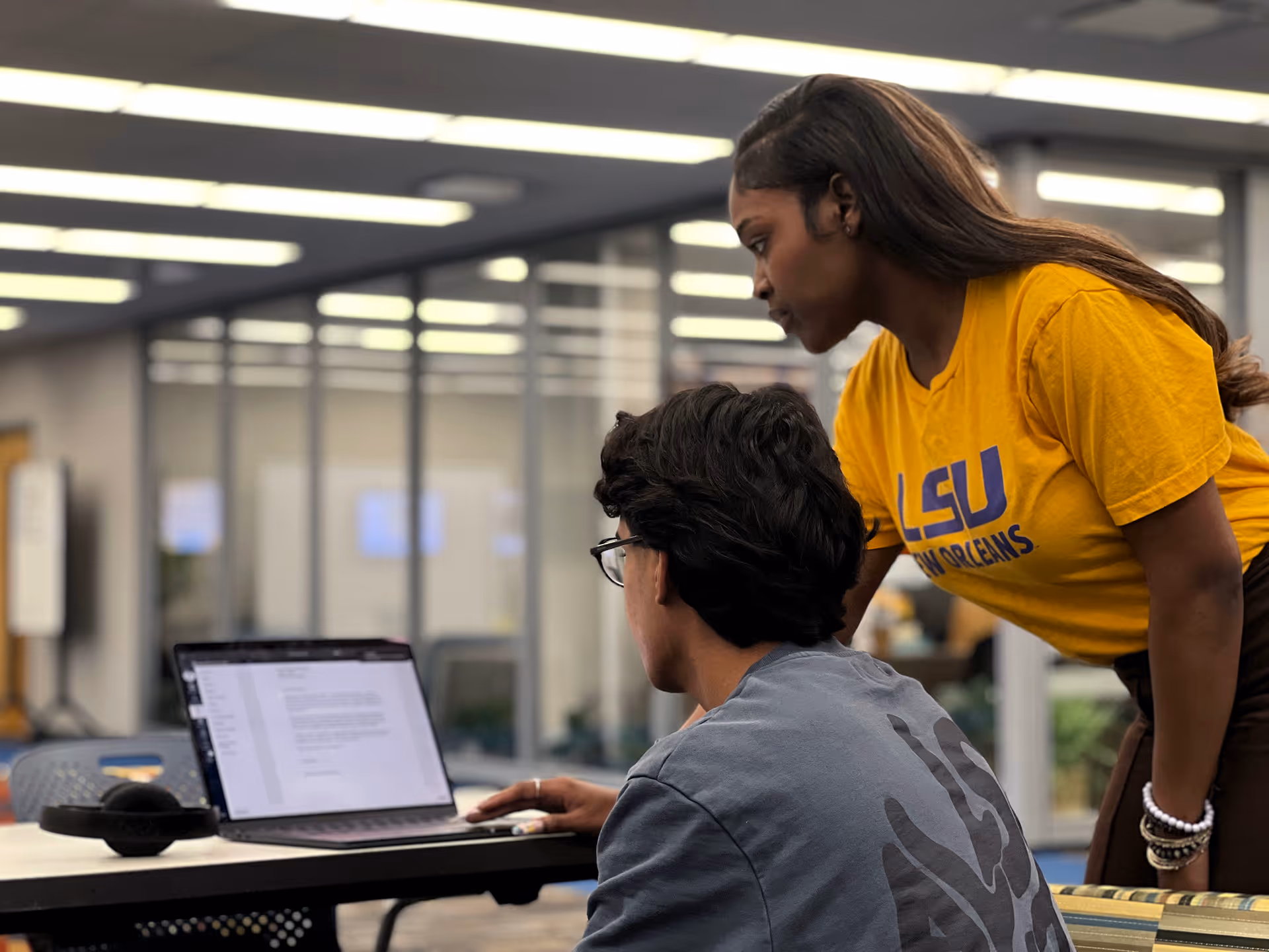 Woman in yellow LSU shirt helping a man with glasses on a laptop in a modern office.