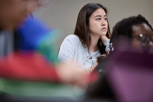Young woman attentively listening in a classroom, surrounded by blurred classmates.