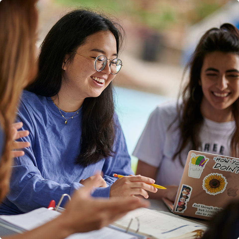 Students smiling and studying together with a laptop and notebooks outdoors.