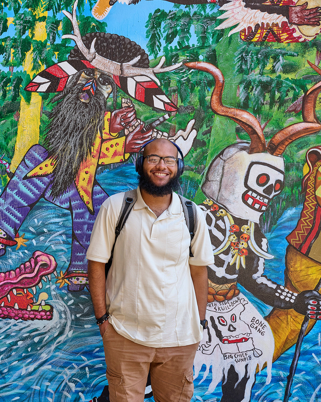 Smiling man with glasses and headphones stands before colorful mural of skeleton and forest figures.