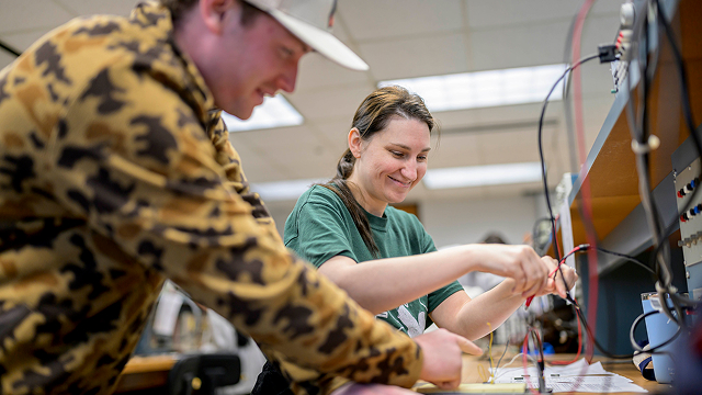 Two people assembling or testing electronic circuits with wires in a classroom or lab setting.