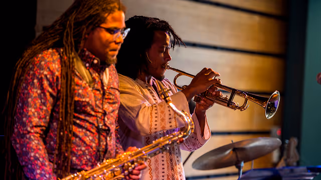 Two musicians playing saxophone and trumpet on stage with wooden wall background.