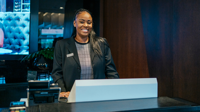 Smiling female hotel receptionist standing behind a front desk in a modern lobby.