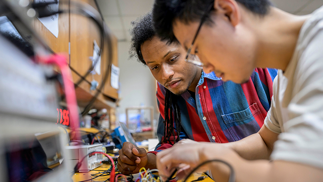 Two students work together on electronics components at a cluttered lab bench.