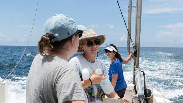 Three women on a boat in the ocean, one holding bottles, under a partly cloudy sky.