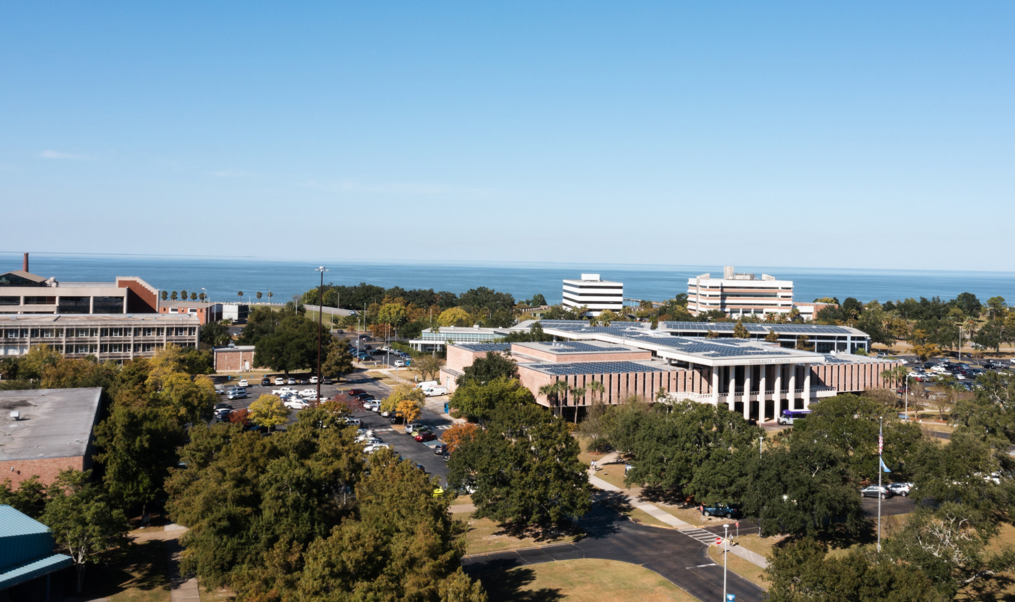 Wide aerial view of LSU New Orleans campus with buildings, parking lots, trees, and a large body of water in the background.