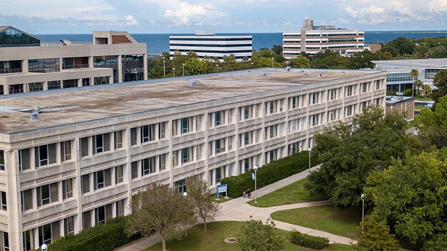 A large multi-story office building surrounded by greenery with ocean visible in the background.