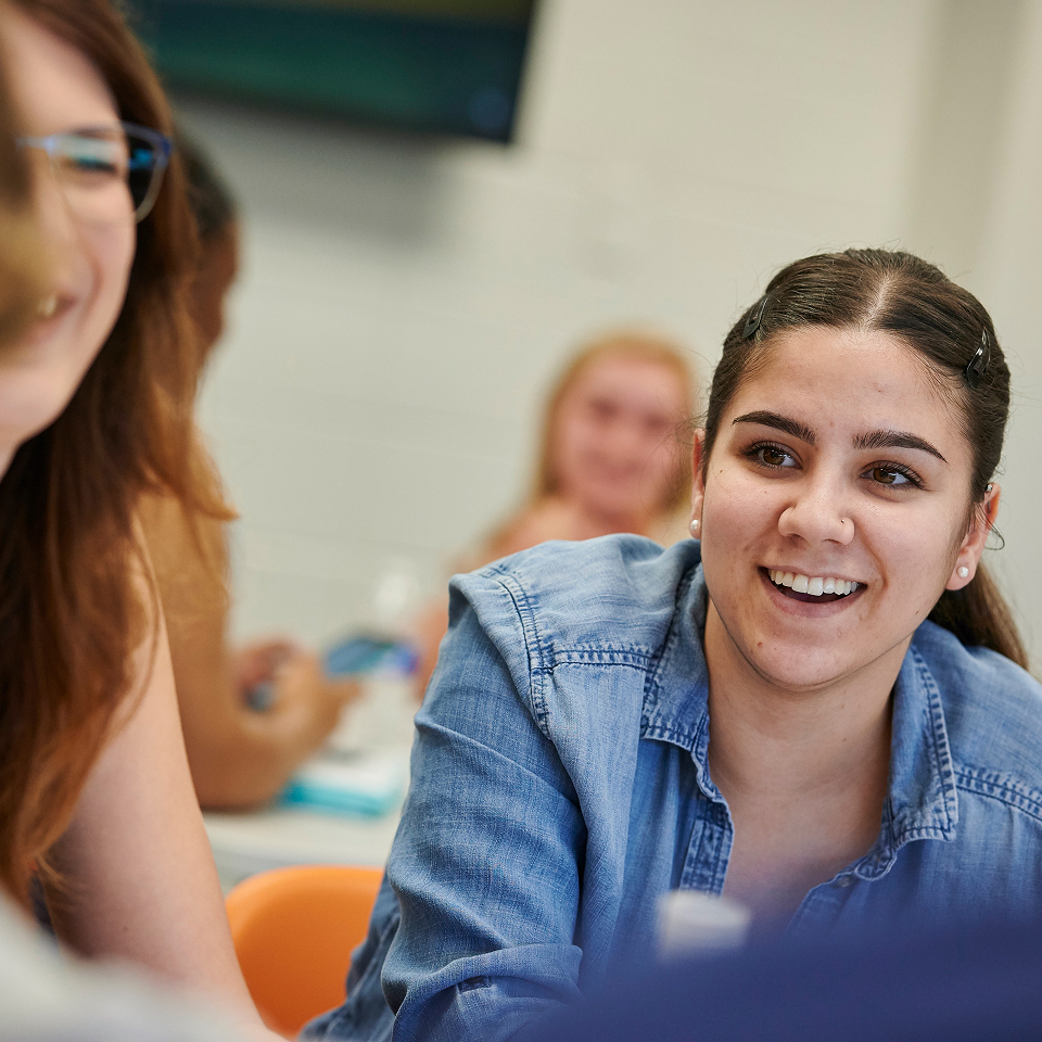 Young woman in a denim shirt smiling and talking with others in a classroom setting.