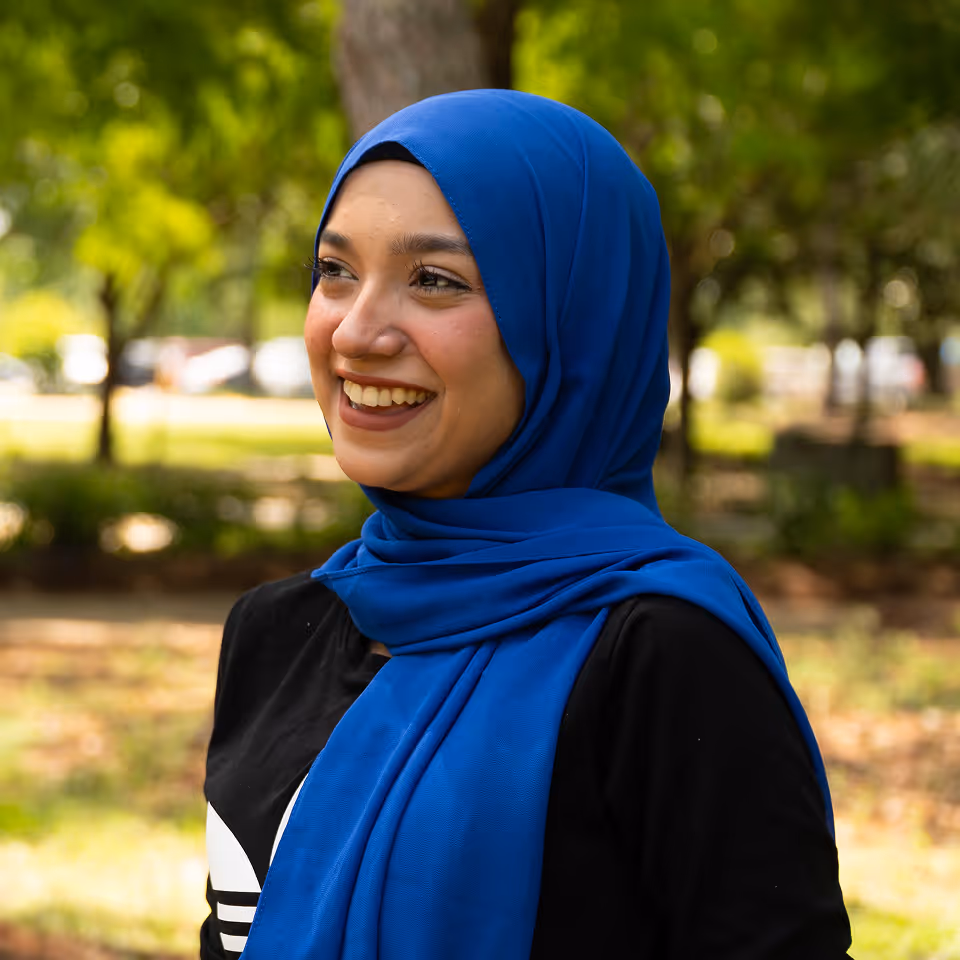 Smiling woman wearing a bright blue hijab and a black top, standing outdoors with greenery in the background.