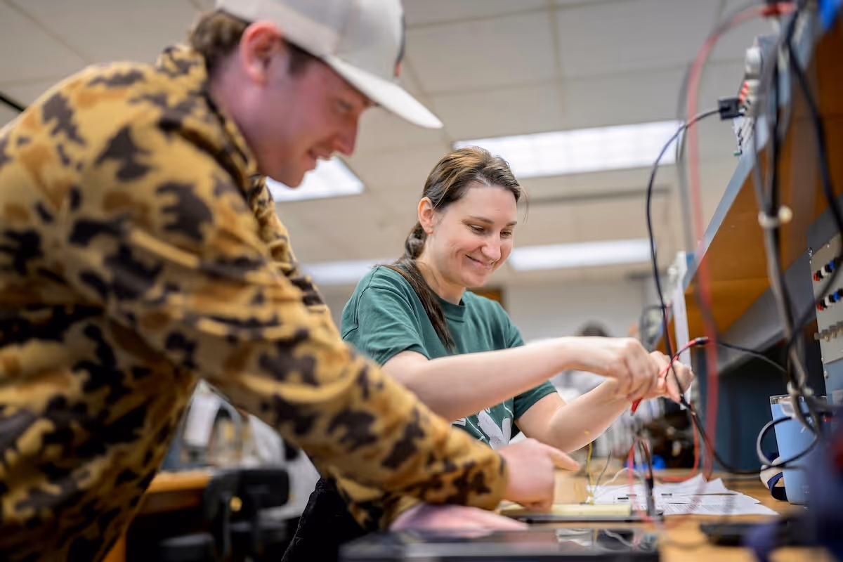 Two adults working together on an electrical wiring project in a lab setting, smiling and focused.